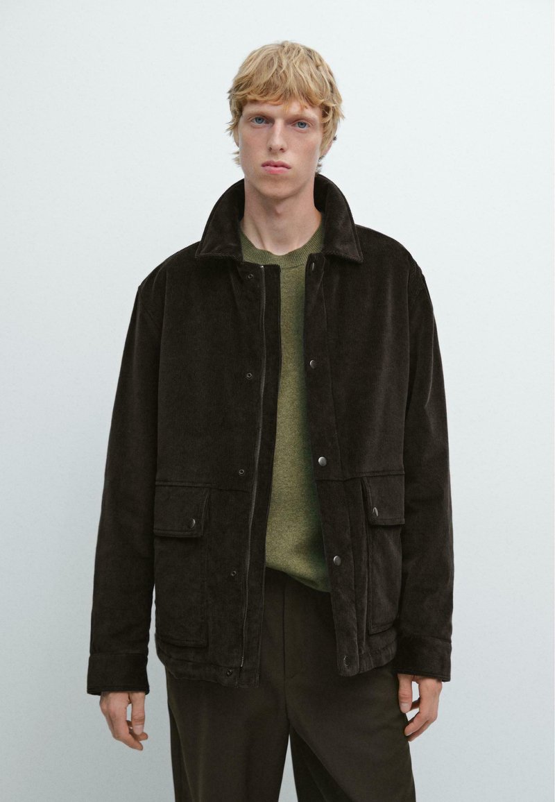 Giovane uomo con capelli biondi che indossa una giacca di velluto a coste marrone scuro su un maglione verde oliva e pantaloni scuri, su uno sfondo chiaro e semplice.