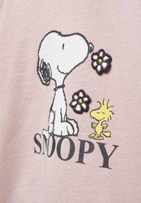 Ljusrosa T-shirt med en grafik av Snoopy och Woodstock, broderade blommor, texturerade detaljer och fet svart text "SNOOPY."