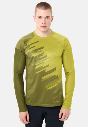 Jeune homme portant un maillot de sport à manches longues vert olive avec des motifs diagonaux abstraits et un pantalon foncé, faisant face à l'avant.