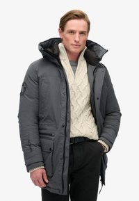 Parka isolée grise avec une fermeture éclair à l'avant, une grande capuche et de multiples poches. Portée sur un pull en grosse maille crémeux clair.