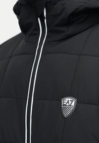 Μαύρο επενδυμένο μπουφάν με κουκούλα, λευκό φερμουάρ και γαζωτό λογότυπο EA7 στο αριστερό στήθος. Η υφή είναι ομαλή με ραμμένα διακοσμητικά στοιχεία.