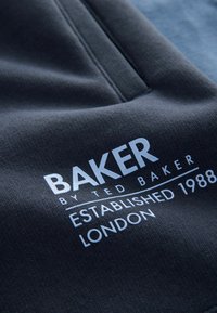Tumši pelēks audums ar drukātu logotipu: "BAKER by TED BAKER," balts teksts, ar "Izveidots 1988" un "LONDONA" zem tā. Gluda tekstūra.