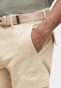 Άτομο που φοράει μπεζ φόρμα με πλαϊνές τσέπες και υφασμάτινη ζώνη, με το χέρι να ακουμπά στην μπροστινή τσέπη μπροστά σε ουδέτερο φόντο.