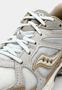 Lys grå sneaker med mesh- og semsket skinnmateriale, hvite snørebånd og et beige akcentdesign med en distinkt logo på tungen.