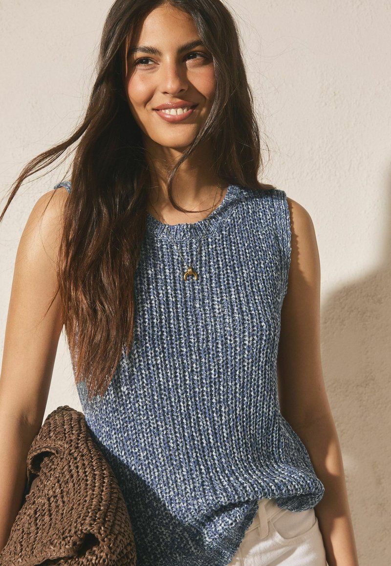 Jeune femme aux longs cheveux bruns portant un haut bleu en maille sans manches et un pantalon blanc, portant un sac tressé marron, souriant légèrement.