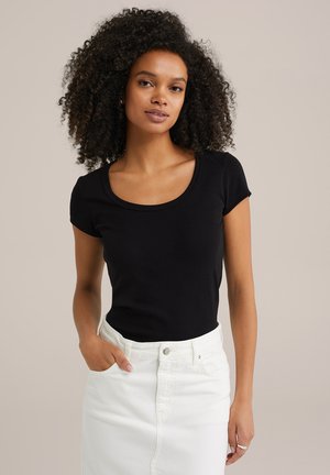 Haut noir à manches courtes avec un col arrondi, fabriqué en tissu doux. Le mannequin le porte avec une jupe en denim blanche, les mains dans les poches.