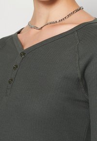 Dunkelgrünes, geripptes Langarmshirt mit V-Ausschnitt, drei schwarzen Knöpfen am Ausschnitt und sichtbaren Nahtakzenten.