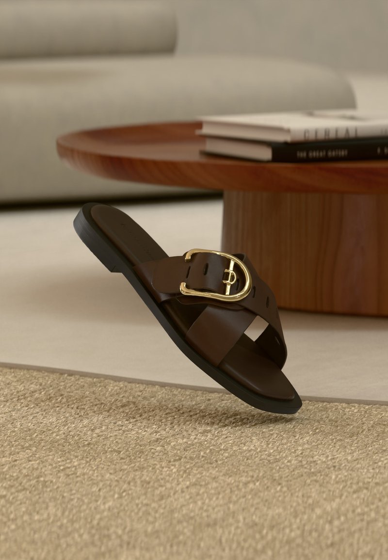 Καφέ δερμάτινο σανδάλι τύπου slip-on με χρυσή αγκράφα πάνω σε μπεζ χαλί, ξύλινο τραπεζάκι καφέ και βιβλία στο φόντο.