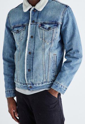 Uomo che indossa una giacca di jeans azzurro chiaro con colletto in sherpa bianco, chiusura frontale con bottoni e pantaloni neri, in piedi davanti a uno sfondo semplice.