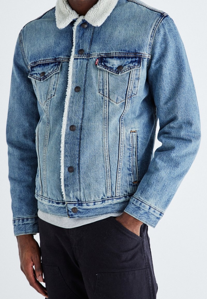 Homme portant une veste en denim bleu clair avec un col en fourrure blanche, une fermeture par boutons à l'avant et un pantalon noir, debout devant un fond uni.