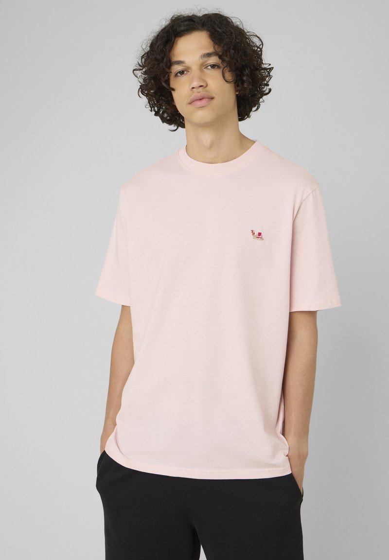 Jeune homme aux cheveux bouclés portant un t-shirt rose clair oversize et un pantalon noir, mains partiellement dans les poches, fond gris neutre.