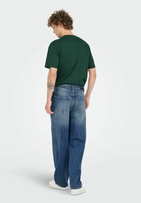 Grön bomullströja med korta �ärmar, kombinerad med lösa blå denimjeans. Outfiten visar en bakvy med synlig sömnad och fickor.