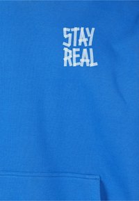 Felpa di cotone blu con grafica bianca testurizzata "STAY REAL" sul petto e tasca a marsupio in basso.