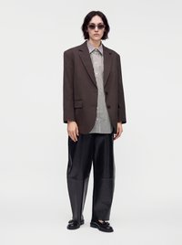 Άτομο που φοράει σκούρο καφέ oversized σακάκι, ριγέ πουκάμισο με κουμπιά, φαρδιά μαύρη παντελόνια, μαύρα παπούτσια και γυαλιά ηλίου, στέκεται μπροστά σε ανοιχτό φόντο.