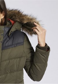 Polstret parka-jakke i olivengrøn med en sort læderakcent på skulderen, aftagelig pelshætte og ribbede manchetter. Har trykknaps- og lynlåslukninger.