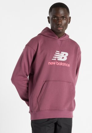 Persoon draagt een mauve New Balance hoodie met voorzak en logo, staand tegen een effen lichte achtergrond, gezicht naar voren gericht.