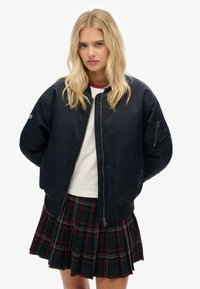 Marine bomberjack met rits, geribbelde manchetten en zakdetails. Draag het over een wit shirt en combineer het met een groene geruite plooienrok.