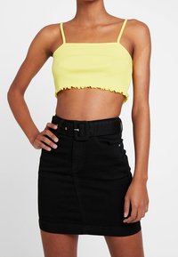 Top corto amarillo con tirantes finos y dobladillo volado, combinado con una falda de mezclilla negra de cintura alta que incluye un cinturón y bolsillos frontales.