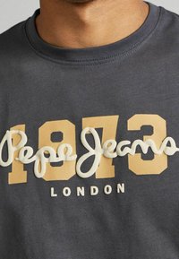 Primer plano de una camisa gris con números beige "1973", texto blanco texturizado "Pepe Jeans" y pequeño texto blanco "London" debajo.