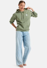 Sudadera verde con un bolsillo frontal y cordones plateados, que presenta un gráfico de sol. Combinada con unos pantalones vaqueros de pierna ancha de color azul claro y sandalias.