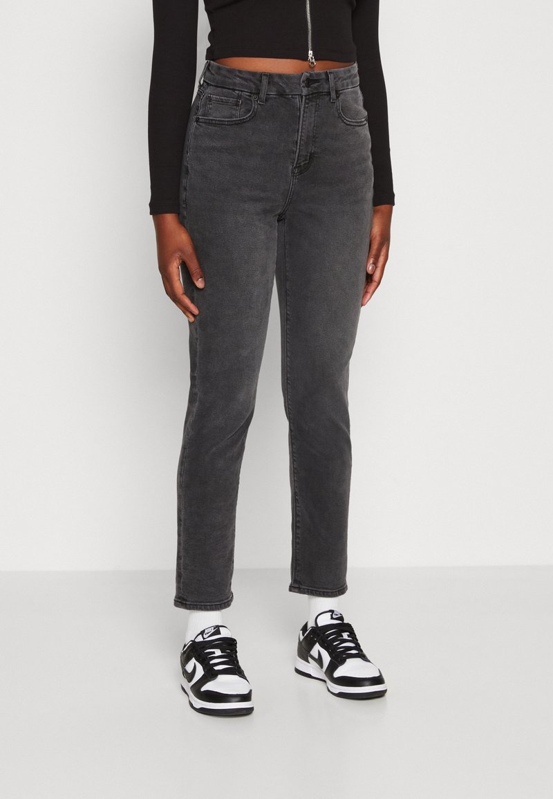 Personne portant un jean noir délavé taille haute, un haut noir court à manches longues, des chaussettes blanches et des baskets noir et blanc, debout contre un mur blanc.