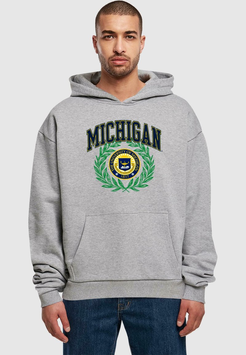 Sudadera gris con la inscripción "MICHIGAN" en texto azul y un emblema circular en verde y amarillo centrado en la parte delantera. Incluye un bolsillo tipo canguro.