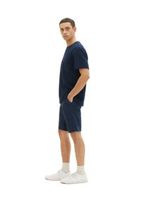 Marineblaues T-Shirt mit kurzen Ärmeln und Brusttasche, kombiniert mit passenden marineblauen Shorts. Als Schuhwerk werden weiße Sneakers mit hellgrauen Socken getragen.