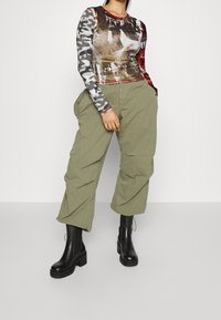 Top met lange mouwen van stof met meerdere patronen in rood, zwart en grijs. Olijfgroene cargobroek en kniehoge zwarte legerlaarzen maken de look compleet.