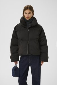 Svart puffjakke med høy krage, teksturert overflate og glidelås foran. Modellen holder en liten marineblå håndveske og har på seg mørke jeans.