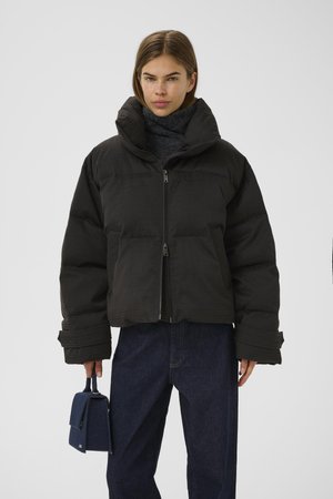 Black puffer jacket with a high collar, textured surface, and front zipper. Model holds a small navy handbag, wearing dark jeans.