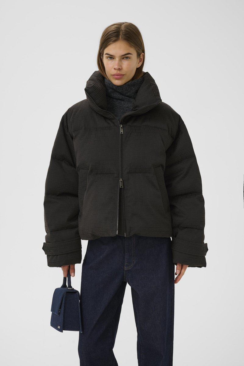 Svart puffjakke med høy krage, teksturert overflate og glidelås foran. Modellen holder en liten marineblå håndveske og har på seg mørke jeans.