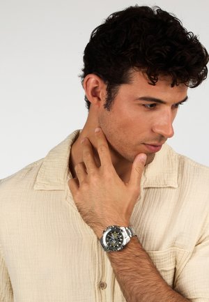 Montre en acier inoxydable argenté avec un cadran noir et des fonctions de chronographe, portée sur un poignet à la peau bronzée, contre une chemise beige clair.