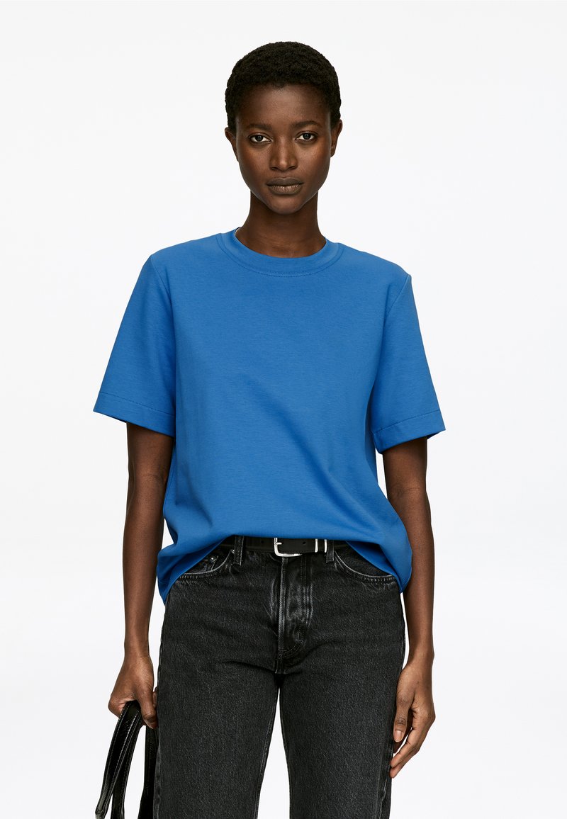 T-shirt bleu à manches courtes en tissu de coton, col rond, coupe décontractée, associé à un jean noir taille haute et tenant un sac noir.