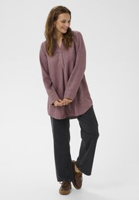 Ženska se smeška in gleda navzdol, oblečena v lila dolgo tuniko, črne široke hlače in rjave čevlje z vezalkami na belem ozadju.