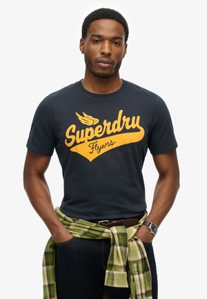 Mand iført navyblå "Superdry Flyers" t-shirt, grøn ternet skjorte bundet om livet, mørke jeans og sort armbåndsur, stående med hænderne i lommerne.