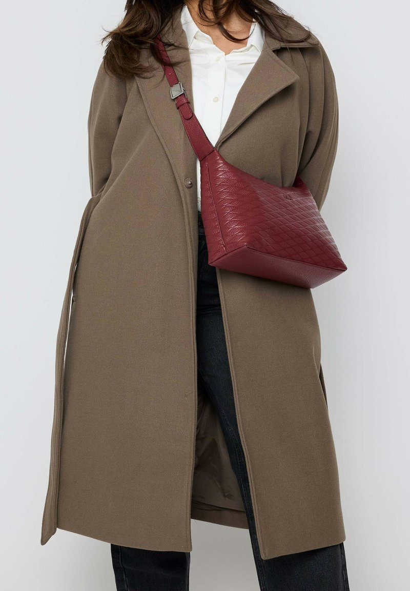 Burgundy textured leather shoulder bag with a triangular shape, complemented by a matching adjustable strap, worn over a taupe coat.