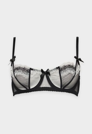 Une femme porte un ensemble de lingerie noire et transparente, bordé de dentelle blanche, sur un fond uni et clair.