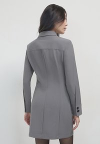 Cappotto grigio su misura con una texture liscia, maniche lunghe e un colletto strutturato. Presenta tre bottoni neri su ciascun polsino e una silhouette aderente.
