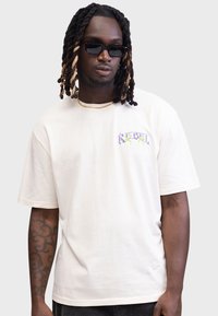 Weißes Baumwoll-T-Shirt mit kurzen Ärmeln, das ein buntes "REBEL"-Design auf der linken Brust zeigt. Glatte Textur, lässige Passform, einfacher Ausschnitt.