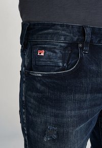Jeans de mezclilla oscura con una textura desgastada y desvaída. Presenta una pequeña etiqueta de logo roja en el bolsillo frontal y detalles de desgaste menores en la tela.