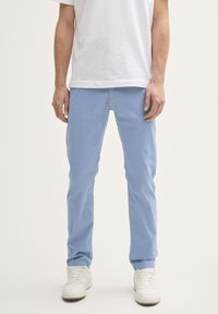 Lichtblauwe katoenen broek met een slim fit, voorzien van zijzakken en een tailleband. Gecombineerd met witte sneakers op een effen achtergrond.