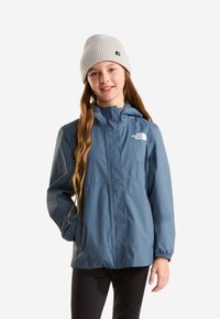 Fille aux longs cheveux portant un bonnet beige, une veste bleue The North Face et un pantalon noir, debout avec une main dans sa poche.