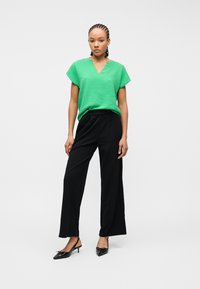 Γυναίκα με πλεξουδάκια που φοράει μια υφασμάτινη πράσινη κοντομάνικη μπλούζα, μαύρο παντελόνι με φαρδιά πόδια και μαύρα παπούτσια στιλέτο με λουράκι, στέκεται όρθια.