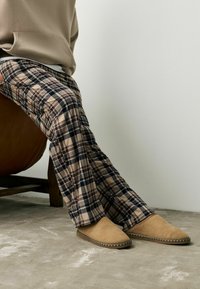 Beige suede slippers with a pointed toe and decorative woven trim, paired with plaid flannel pants in beige and navy tones.
