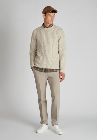 Giovane uomo con berretto marrone, maglione di maglia beige sopra una camicia a quadri, pantaloni beige e sneakers bianche in piedi davanti a uno sfondo semplice.