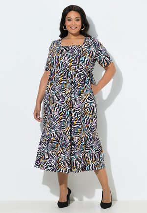 Kobieta stoi, ubrana w kolorową midi sukienkę w paski zebry z krótkimi rękawami i czarne szpilki, uśmiecha się na tle jednolitego tła.