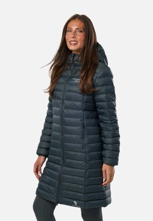 Femme portant un long manteau d'hiver matelassé à capuche de couleur sombre, debout devant un fond clair uni.