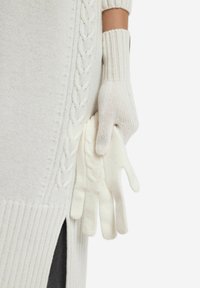 Białe, dziane rękawiczki z prążkowanym mankietem, z detalami w stylu warkocza. Miękka tekstura, trzymane razem z dużym białym swetrem w swobodnej pozycji.
