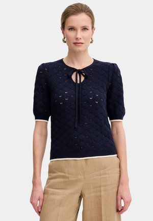 Donna che indossa una camicetta blu navy testurizzata a maniche corte con dettaglio annodato al collo e pantaloni beige a vita alta, in piedi davanti a uno sfondo semplice.