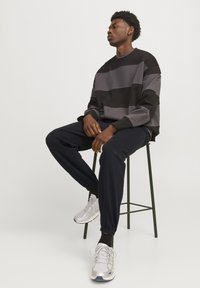 Grijs en zwart gestreepte sweatshirt, relaxte pasvorm, gecombineerd met donkere joggers en grijze sneakers, zittend op een groene metalen kruk.